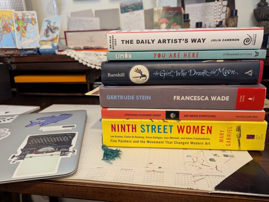 A stack of books on a cluttered desk. From top to bottom: The Daily Artist's Way by Julia Cameron; You Are Here edited by Ada Limon; The Girl Who Drank the Moon by Kelly Barnhill, Gertrude Stein by Francesca Wade; Art Above Everything by Stephanie Elizondo Griest; and Ninth Streeet Women by Mary Gabriel. Books to the right of a silver laptop covered with stickers, and atop a desk calendar mat covered with various colored ink scribbles.